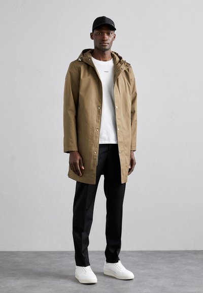 Impermeabile khaki con cappuccio, bottoni a scatto e tasche frontali, abbinato a una maglietta bianca, pantaloni neri e sneakers bianche. Vestibilità casual.
