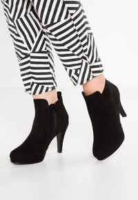 Černé semišové kotníkové boty s elegantním designem, špičatou špičkou a bočním zipem. Nosené s černobílými pruhovanými kalhotami.