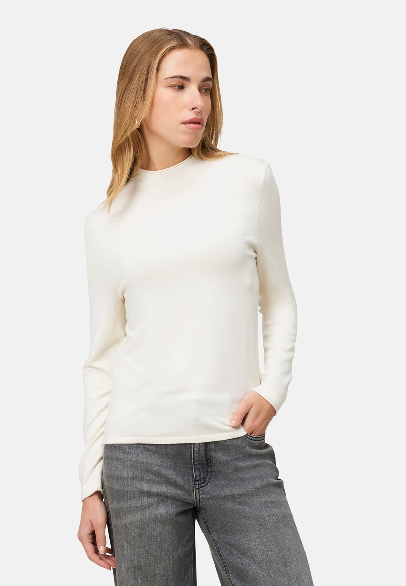 Pull blanc à manches longues avec col rond en tissu doux, présentant une coupe ajustée et une texture lisse, associé à un jean en denim gris.