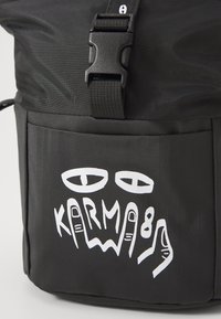 Sac à dos noir en tissu texturé avec un design graphique blanc saisissant représentant des yeux et des doigts, étiqueté "KARMA88".