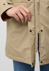 Veste d'extérieur beige avec une fermeture éclair à l'avant, des boutons-pression et une texture lisse. Présente une main près de la fermeture éclair, mettant en valeur le design.