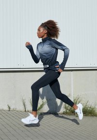 Długi rękaw, zapinana na zamek sportowa koszulka w gradientowym szarym kolorze, w zestawieniu z czarnymi legginsami. Białe buty sportowe z logo. Biegacz w ruchu na utwardzonej nawierzchni.