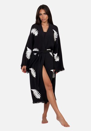 Vrouw die een zwarte kimono-stijl badjas draagt met witte bladmotieven en een gestrikte tailleband, staat blootsvoets met één been iets naar voren.