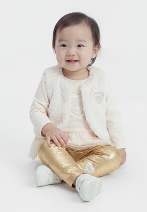 Petite fille assise en tailleur portant une veste blanche en fausse fourrure, une chemise crème, un legging doré brillant et des chaussures blanches sur fond blanc.