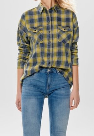 Žena v dlouhém žluto-modrém károvaném košili s knoflíky, která je zastrčená do vysokých modrých džínů, stojí na jednobarevném pozadí.