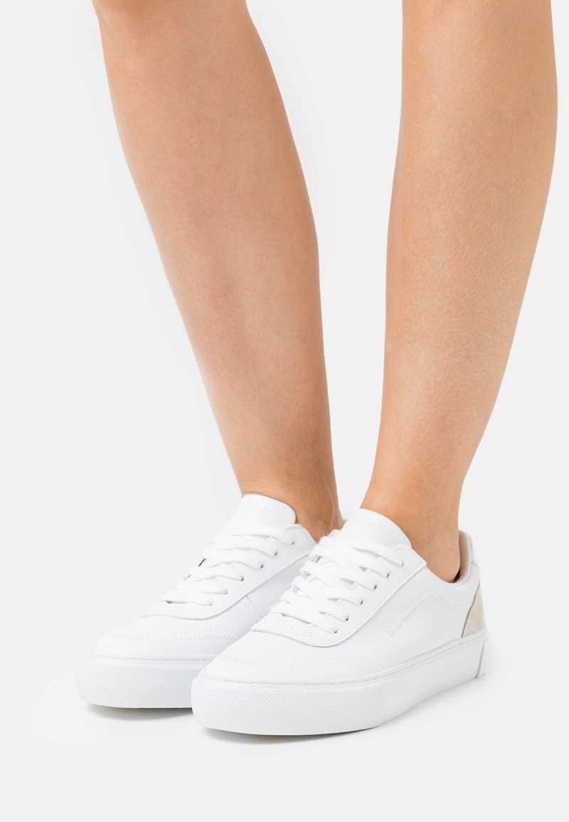 Vita låga sneakers med snörning på ett par bara ben mot en enkel vit bakgrund.