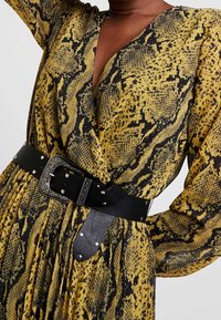 Femme portant une robe imprimée serpent jaune et noire avec des manches longues, cintrée à la taille par une large ceinture noire avec une boucle en argent détaillée.