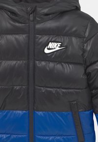 Kurtka puchowa Nike z czarną górną częścią i niebieską dolną częścią. Wykonana z błyszczącego, pikowanego materiału z zamkiem z przodu oraz detalem z logo.