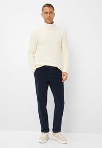 Maglione a collo alto a coste color crema abbinato a pantaloni in corderoy blu navy e sneakers color crema. La trama è morbida e il design è aderente.
