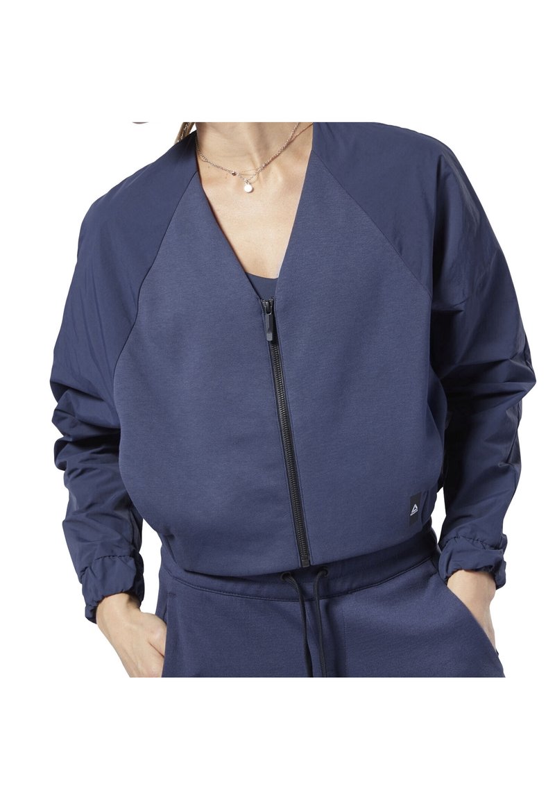 Mörkblå zip-up jacka med V-halsdesign, raglanärmar, elastiska ärmslut och en liten logotyp på fållen. Slät tygkvalitet.