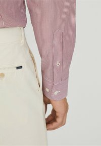 Chemise rayée rouge et blanche avec poignets boutonnés, associée à un pantalon beige avec une étiquette logo. Tissu en coton à texture lisse.
