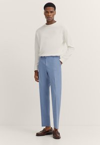 Helleblaue maßgeschneiderte Hose mit glatter Textur, getragen mit einem weißen Langarmhemd, kombiniert mit dunkelbraunen Slippern.