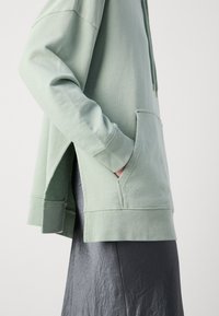Lichtgroene hoodie met een voorzak, zijsplitsingen en lange mouwen, gemaakt van een zachte stof met een gladde textuur.