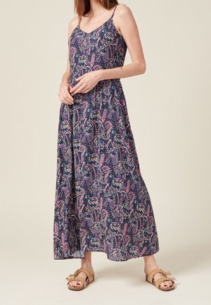 Femme portant une robe sans manches, longue jusqu'aux chevilles, avec un motif cachemire coloré et des sandales marron à enfiler sur un fond uni.