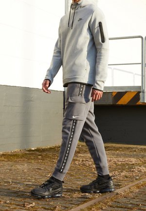Personne marchant en extérieur portant un sweat à capuche Nike gris, un pantalon cargo assorti avec un détail à rayures et des baskets Nike noires sur un sol pavé.