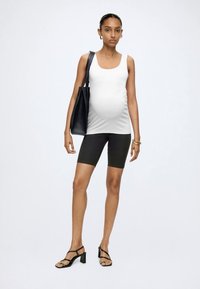 Gravid kvinde iført en hvid tanktop, sorte cykelshorts, sorte sandaler med hæl, og bærer en sort tote-taske over skulderen.