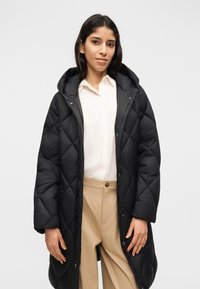 Nainen pukeutuneena pitkään mustaan tikattuun takkiin, jossa on huppu, valkoiseen nappikauluspaitaan ja beigeruskeisiin housuihin, seisoo yksinkertaisen vaalean taustan edessä.