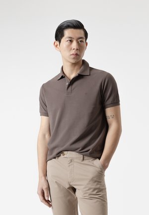 Hombre con camiseta polo marrón y pantalones beige de pie con una mano en el bolsillo, mirando ligeramente hacia un lado contra un fondo liso.