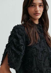 Camicia nera con motivo floreale ricamato, maniche corte, tessuto testurizzato e chiusura con bottoni. Presenta un colletto alto e una vestibilità comoda.