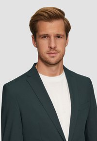 Dunkelgrüner Blazer mit einem einzelnen, geschlitzten Revers, kombiniert mit einem weißen T-Shirt. Verfügt über eine feine Textur und ein Detail in der Brusttasche.