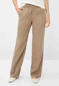 Hellbraune, weit geschnittene Hose mit elastischem Bund, seitlichen Taschen und einem glatten, leichten Stoff. Getragen mit off-weißen flachen Schuhen.