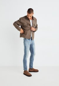Camisa de felpa marrón con botones y dos bolsillos en el pecho, llevada sobre una camisa blanca, combinada con vaqueros ajustados de un azul claro y botas chelsea marrones.