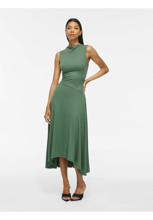 Femme portant une robe midi asymétrique verte sans manches avec un col haut et des talons ouverts noirs, debout devant un fond uni.