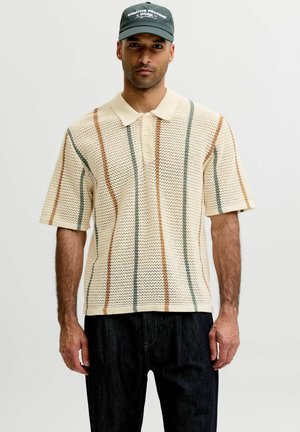 Homme portant un polo en maille crème avec des rayures verticales marron et vertes, une casquette noire avec un texte blanc, et un pantalon foncé, debout devant un fond uni.