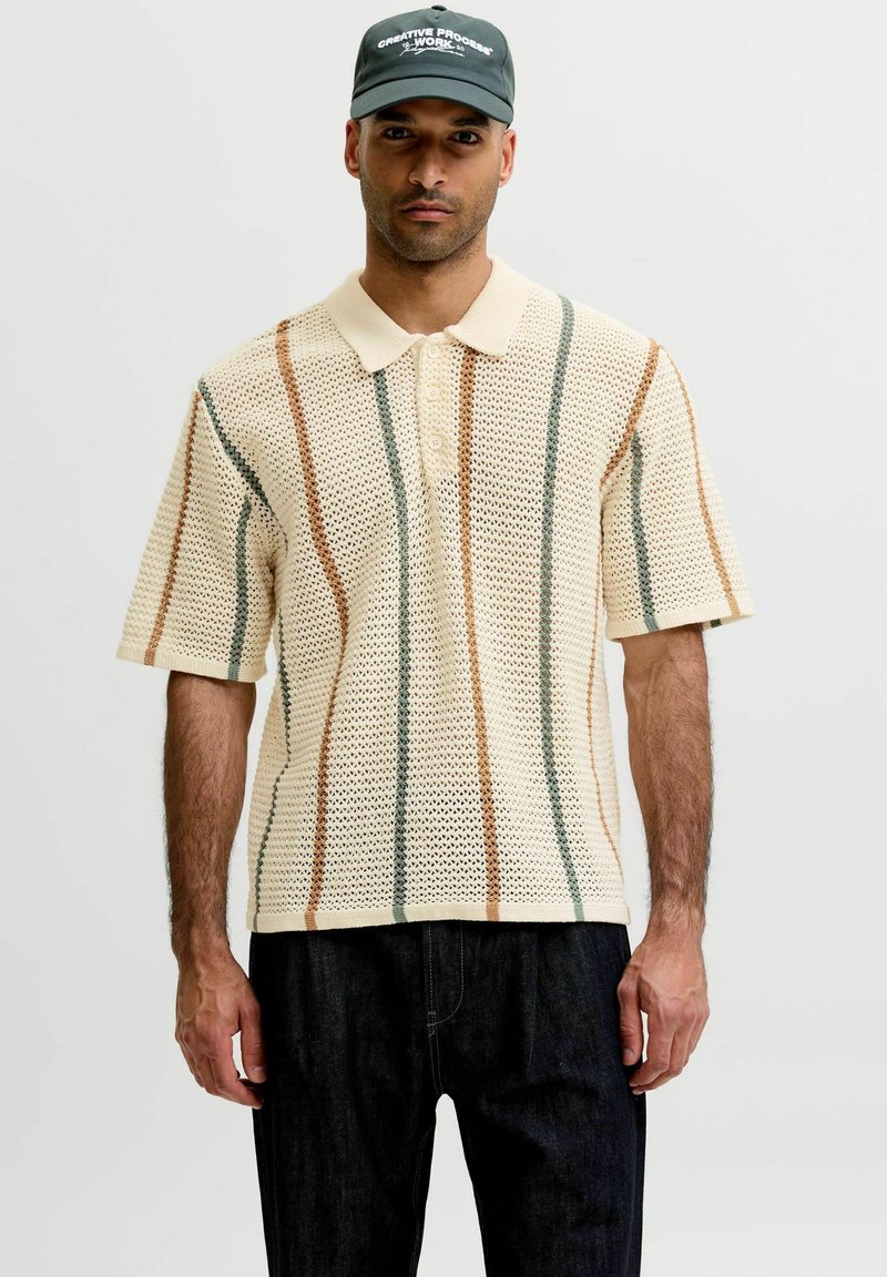 Homme portant un polo en maille crème avec des rayures verticales marron et vertes, une casquette noire avec un texte blanc, et un pantalon foncé, debout devant un fond uni.
