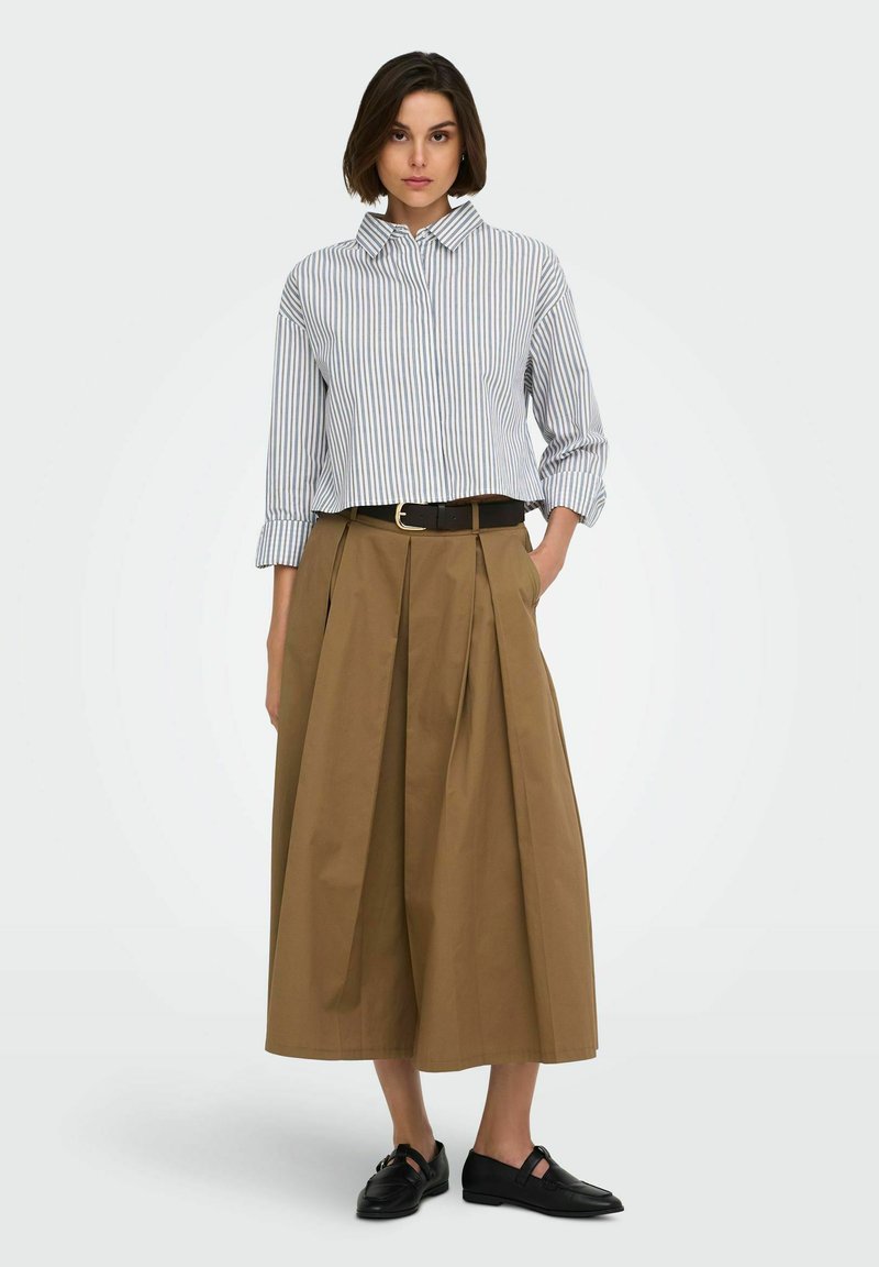 Femme aux cheveux courts et foncés, portant une chemise rayée à manches longues, une jupe large marron taille haute, une ceinture noire et des chaussures plates noires, debout sur fond blanc.