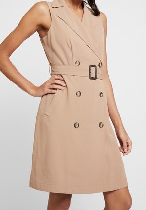 Mouwloze beige trenchcoatjurk met dubbele knopen en tailleceintuur, gedragen door een vrouw met hand op de heup tegen een effen achtergrond.