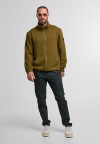 Mann in brauner gemusterter Zip-Jacke, schwarzen Hosen, weißen Sneakers und Sonnenbrille steht vor einem schlichten grauen Hintergrund.