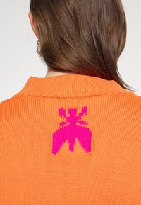 Kirkkaan oranssi neulepusero, jossa on ribattu kaulus ja selässä suuri vaaleanpunainen graafinen kuvio, joka esittää tyyliteltyä hyönteistä.