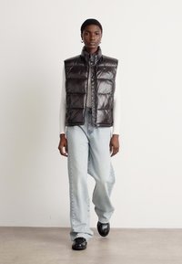 Crni, quilted pufasti prsluk s visokim ovratnikom, nosi se preko bijele majice dugih rukava, u kombinaciji s svijetloplavim hlačama širokih nogavica i crnim cipelama.