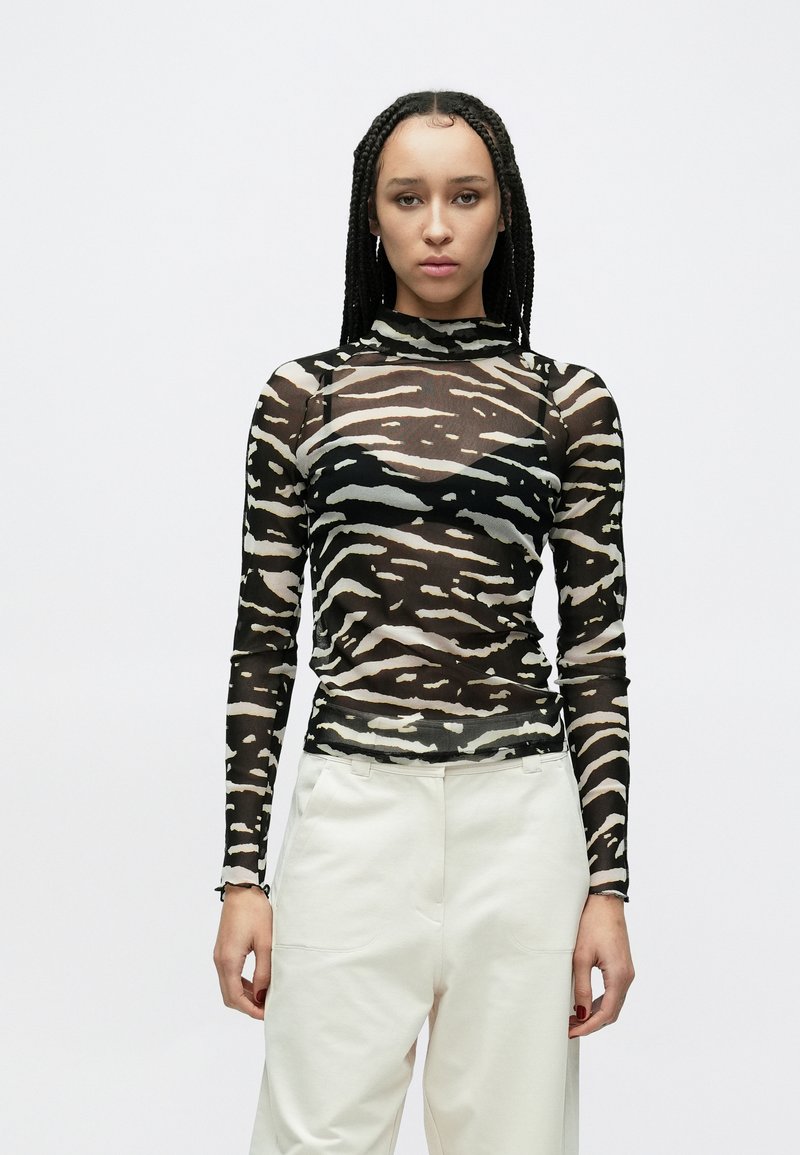 Langærmet, gennemsigtigt top med sort og beige tigerstribemønster. Høj halsudskæring, tætsiddende design og tekstureret stof. Kombineret med hvide bukser.