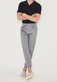 Černé polo tričko s krátkým rukávem a dvěma knoflíky, šedé vzorované kalhoty, a béžové slip-on boty s tlustou bílou podrážkou.