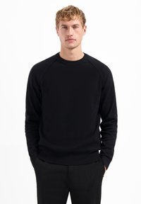 Schwarzer Strickpullover mit geripptem Muster, Rundhals-Auschnitt und Raglanärmeln. Kombiniert mit einer schwarzen Hose, vor einem weißen Hintergrund.