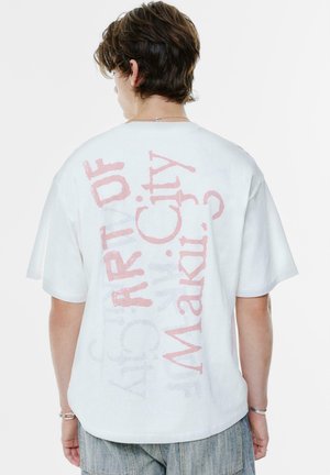 Ung person med kort brunt hår, iført en hvid oversized T-shirt med pink tekst "ART OF Maaki. City ?" på ryggen og denimbukser.