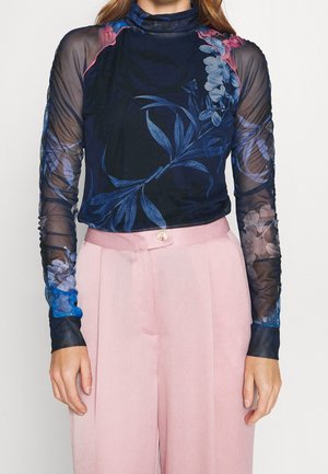 Vrouw draagt een doorschijnende marineblauwe top met lange mouwen met blauwe en roze bloemmotieven en lichtroze hoge taille broek met knoopsluiting.