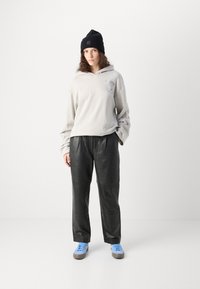 Felpa grigia chiara con un design grafico, pantaloni in pelle nera e sneaker blu. Il modello indossa un berretto nero. Texture morbide e vestibilità comoda.
