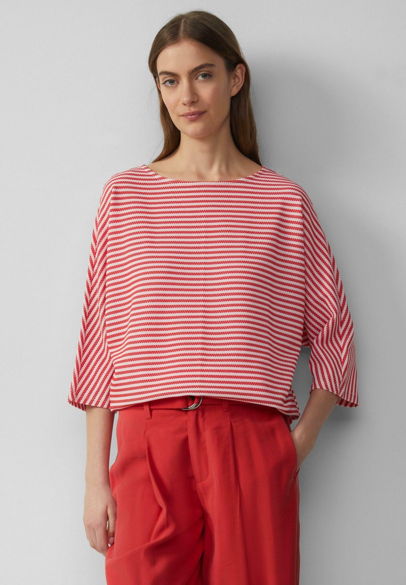 Rot und weiß gestreiftes Strickshirt mit weitem Rundhalsausschnitt und Dreiviertelärmeln, kombiniert mit roten Hosen. Sanfte Textur, entspannte Passform.