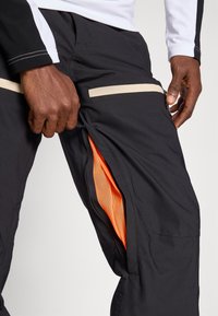 Fekete kültéri nadrág, cipzáras oldalzsebbel, narancssárga hálós béléssel és bézs kiegészítőkkel, amely kiemeli a funkcionális és letisztult formatervezést.