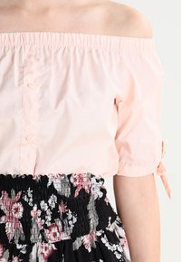 Helle rosa schulterfreie Bluse mit kurzen Ärmeln, mit einem Knopfverschluss vorne. Kombiniert mit einem schwarzen, blumigen, gerafften Rock.