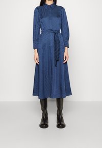 Blå sidentklänning med krage, långa ärmar och banden i midjan, prydd med en strukturerad diamantmönster. Buren med knähöga svarta stövlar.