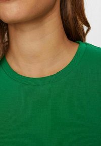 Zbliżenie na szyję osoby i brązowe włosy, noszącej zielony, prążkowany t-shirt z okrągłym dekoltem na jednolitym tle.