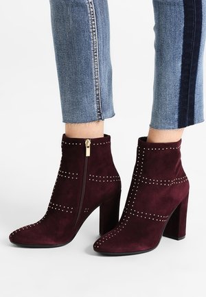 Dunkelburgunderfarbene Stiefeletten aus Wildleder mit Blockabsätzen, genietetem Schmuck, seitlichem Reißverschluss, getragen zu gekürzten blauen Jeans.