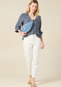 Camicia a fantasia blu, pantaloni bianchi e borsa a tracolla blu con logo. L'outfit è completato da sandali beige con fibbie.