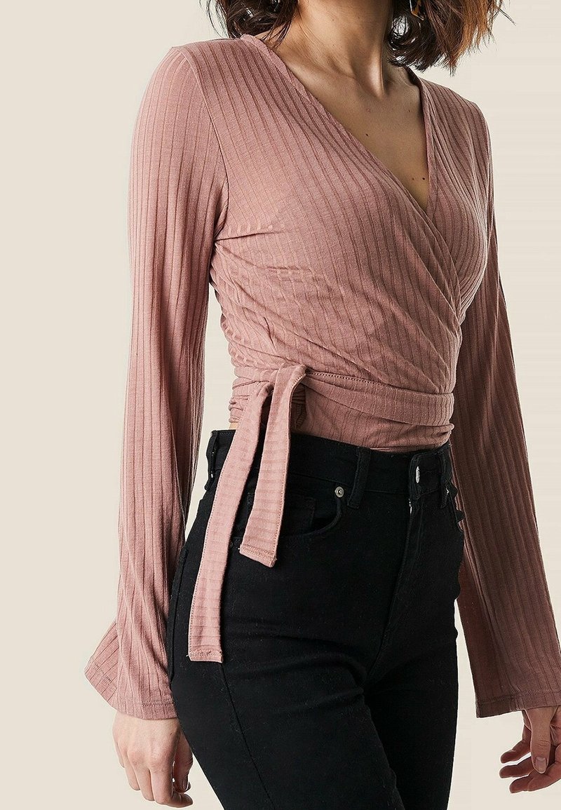 Långärmad, dammig rosa omslagstopp i ribbat tyg, med en djup V-ringning och en knytning i midjan, ihop med svarta jeans.