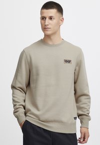 Sudadera beige de algodón suave, con cuello redondo, puños acanalados y una pequeña etiqueta en el pecho. Acabado de tela texturizada.