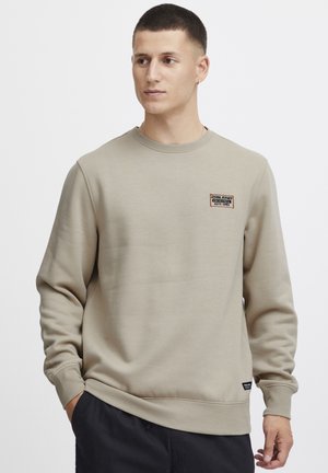 Beige sweatshirt made of soft cotton, featuring a crew neck, ribbed cuffs, and a small label patch on the chest. Textured fabric finish.